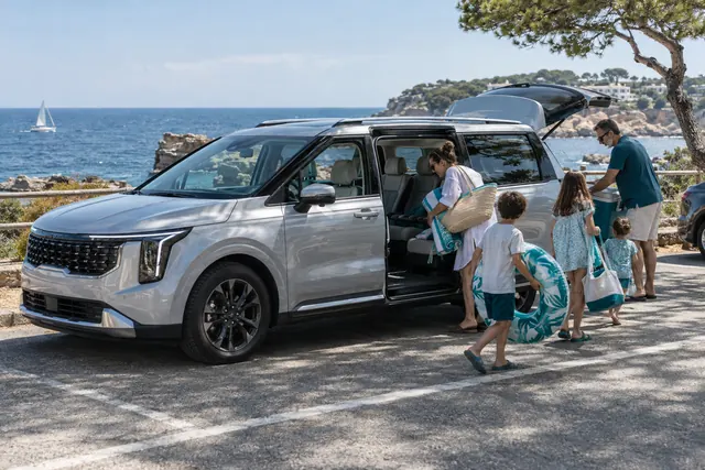 Kia Carnival: большая семья выгружает пляжные вещи из серебристого минивэна с открытыми сдвижными дверями